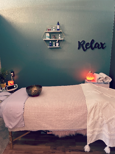 The massage table dressed in cream linens beneath a Relax sign and honeycomb shelves